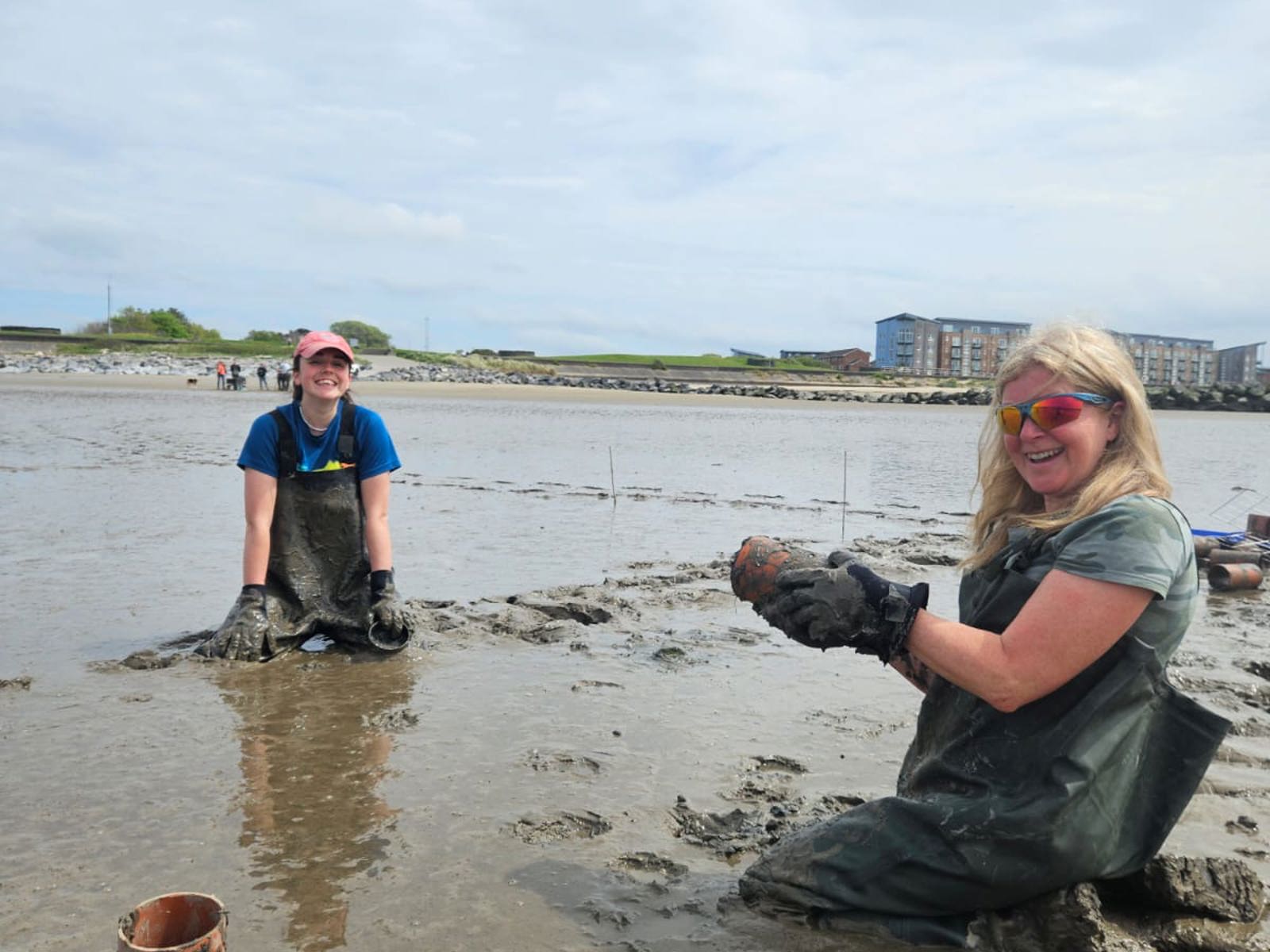 [Carmarthen Bay & Estuaries European Marine Site Officer Judith and Project Seagrass South and West Wales Project Lead Emma are wearing waders and kneeling in muddy sediment at Llanelli beach as they collecting cores.] 