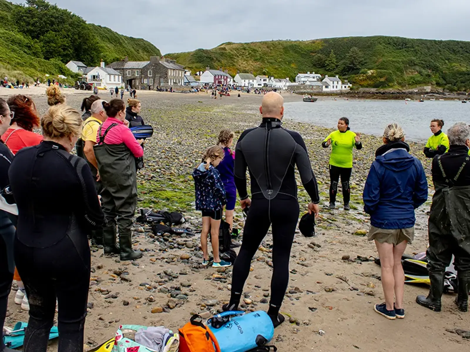 [Volunteers are gathered in a group on the beach at Porthdinllaen listening to North Wales Project Support Amel deliver a safety briefing ahead of a volunteer event. In the background are the buildings of Porthdinllaen bay.