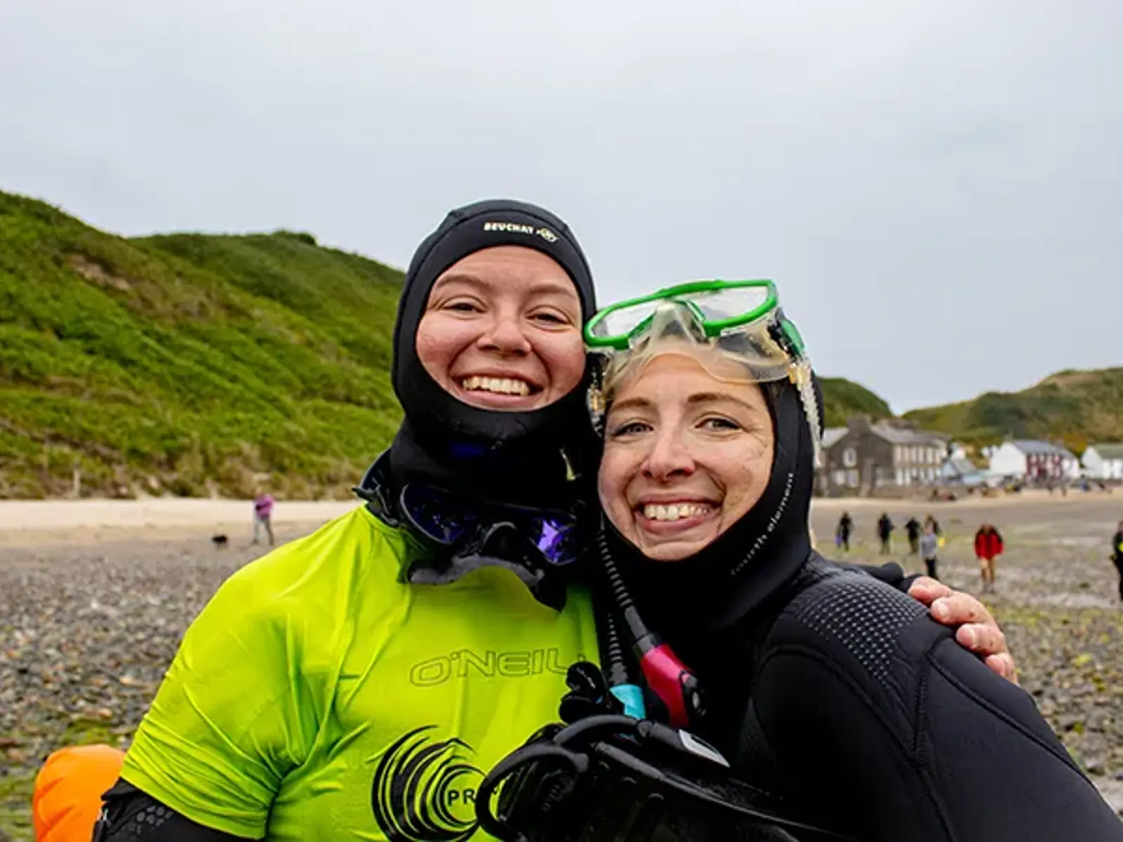 [ One of Project Seagrass' interns is standing on the beach wearing a drysuit (with cap) and a fluorescent yellow Project Seagrass rash vest. They have their arm around a regular volunteer who is wearing a wetsuit and has a snorkel mask pushed up on their head] 